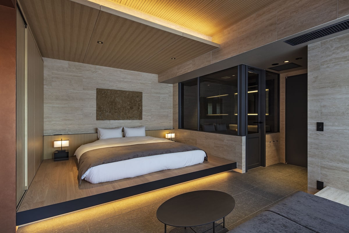 Un dormitorio moderno con una cama grande, lámparas de noche, una mesa redonda y vista al baño a través de una puerta de vidrio.