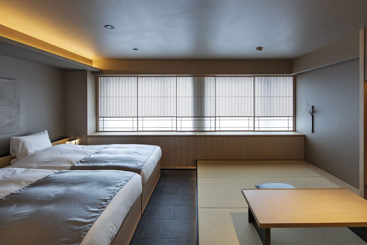 Una habitación de hotel moderna con dos camas, una mesa de madera, esteras de tatami y ventanas amplias con persianas verticales.