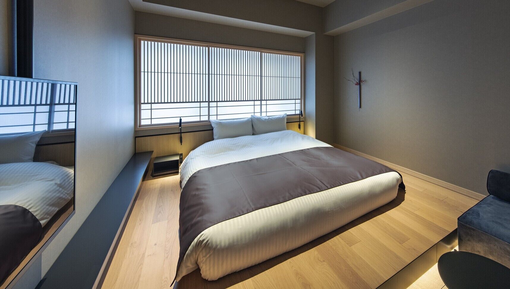 A modern bedroom with a bed, a window with blinds, a bedside table, and a small seating area.