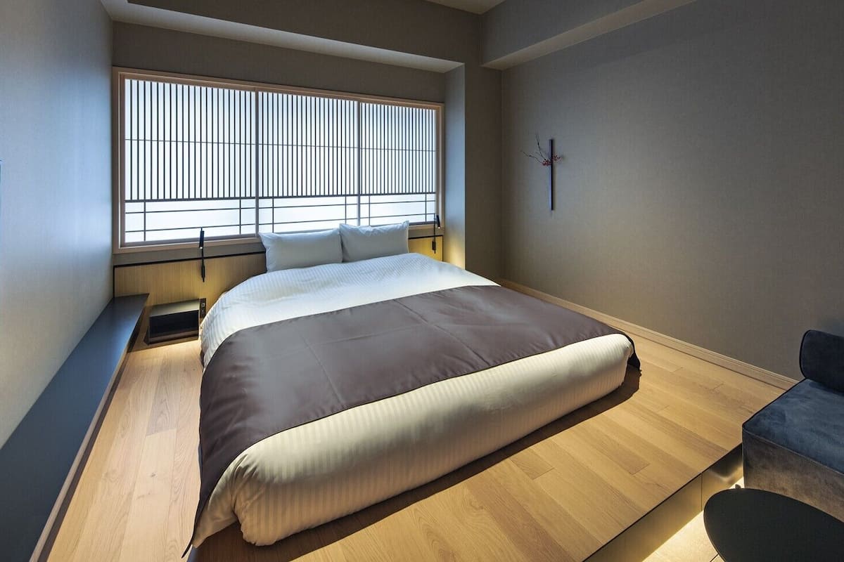 Un dormitorio moderno con cama, ventana con persianas, mesita de noche y una pequeña zona de estar.