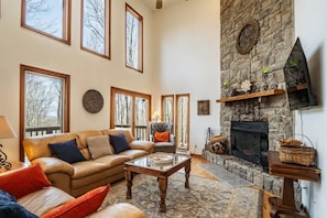 A spacious living room with a stone fireplace, large windows, and a leather sofa. - Timberly - Hot Tub, Dog Friendly, Lake Privileges (Davis)