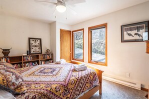 A bedroom with a large bed, a bookshelf, a ceiling fan, and two windows with a view of trees.