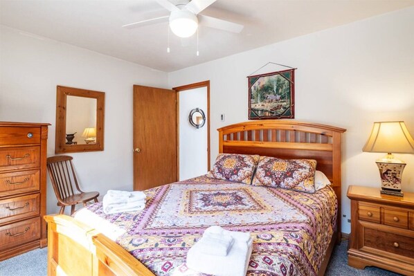 A bedroom with a wooden bed, a nightstand, a chair, a dresser, a ceiling fan, and a wall-mounted mirror.