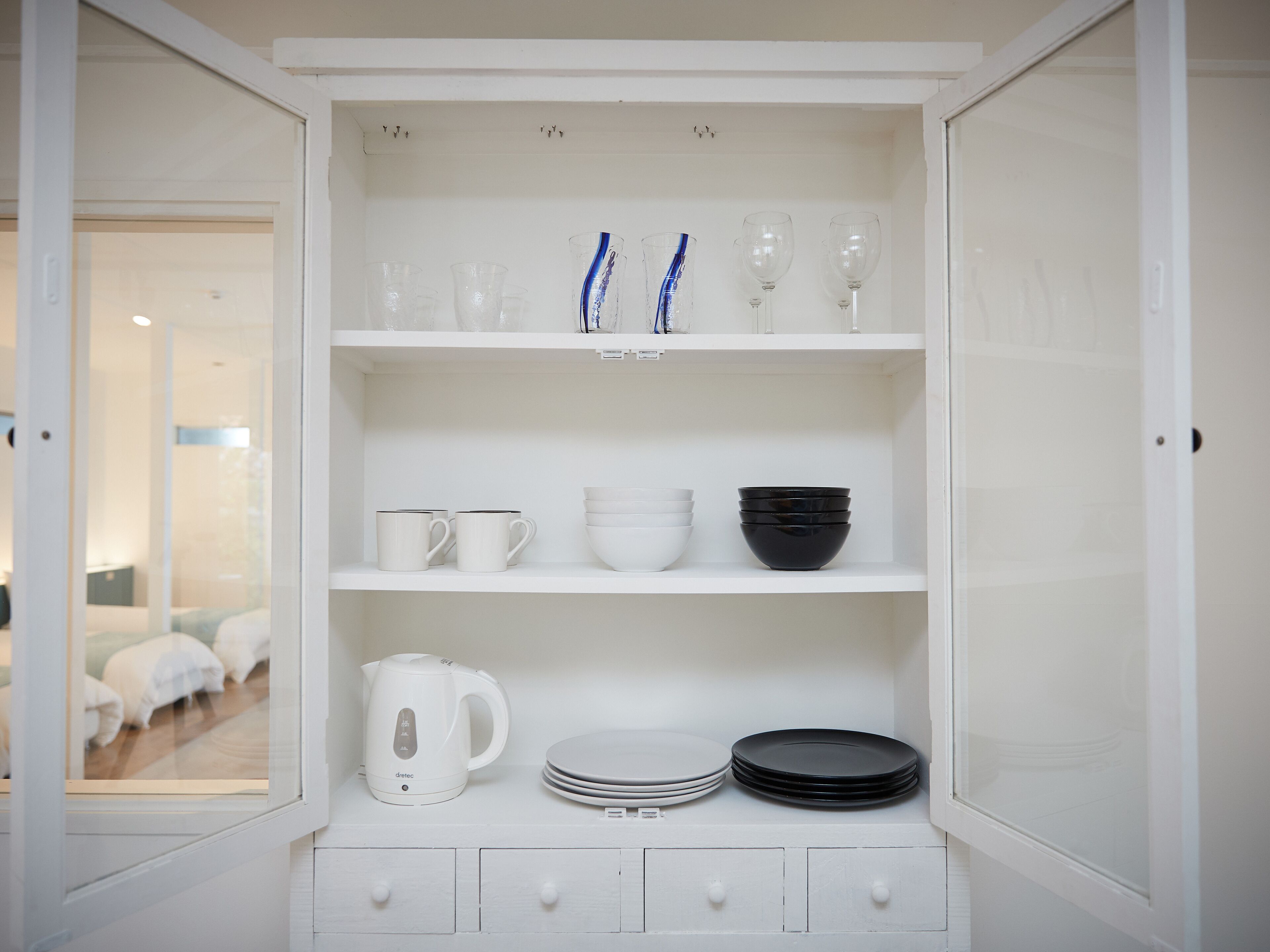 A white cabinet with shelves containing white mugs, bowls, a kettle, and plates.