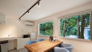 A compact kitchen with a wooden table, blue chairs, and a large window offering a view of trees. - Adan Resort EN (Motobu)
