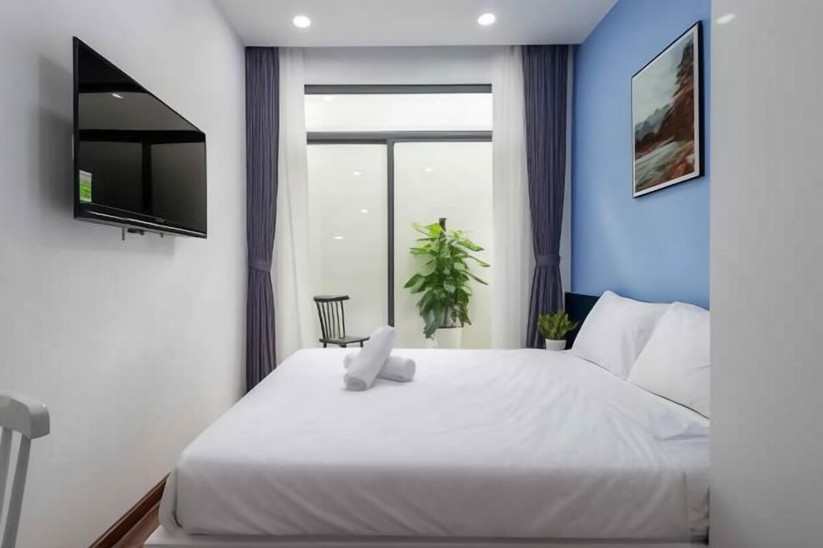 A modern hotel room with a bed, a flat-screen TV mounted on the wall, a window with curtains, a potted plant, and a painting on the wall.