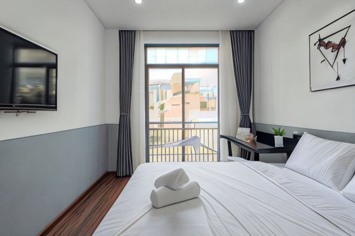 A hotel room with a bed, a TV, a balcony with curtains, and a desk with a lamp.