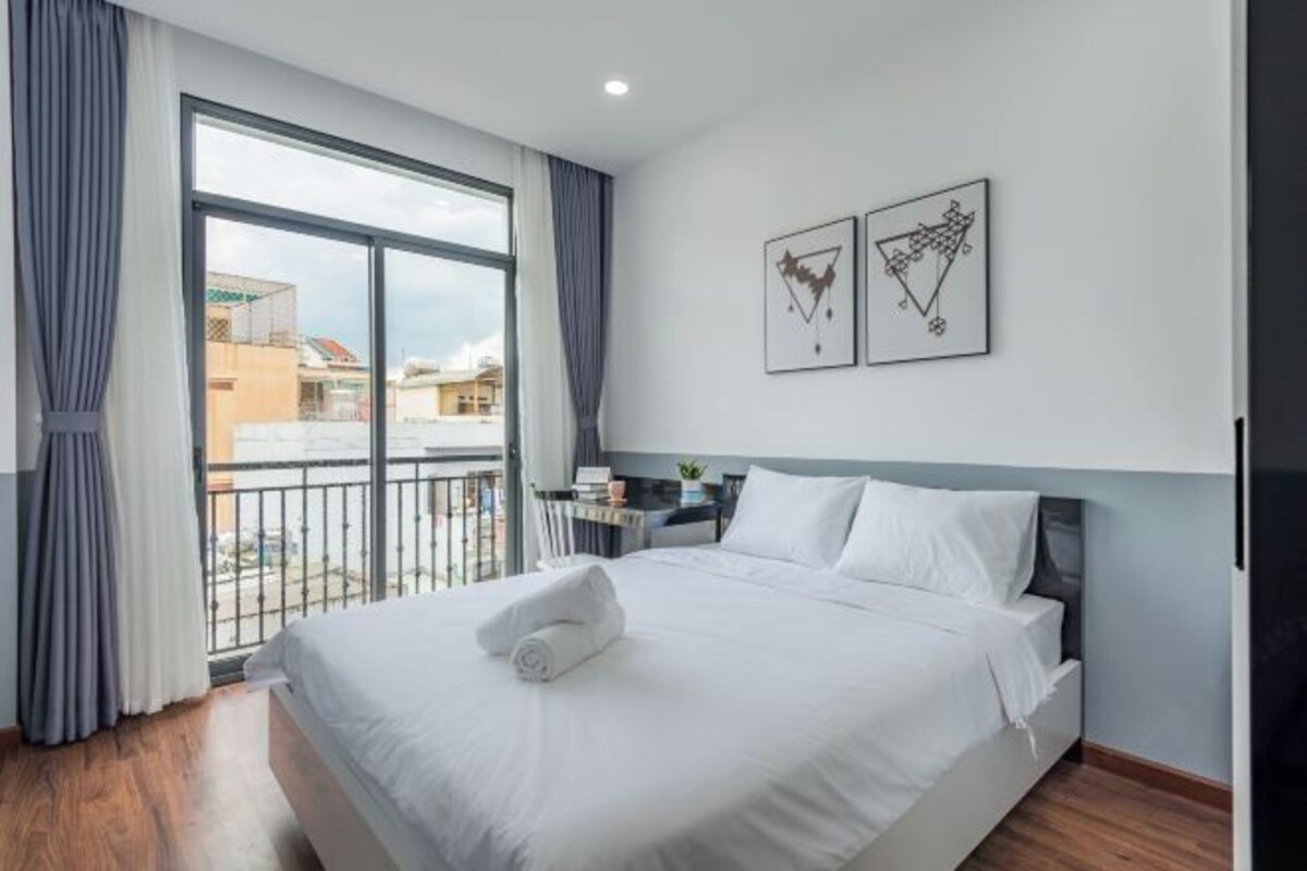A hotel room with a large bed, a view of the city through a balcony, and two framed artworks on the wall.
