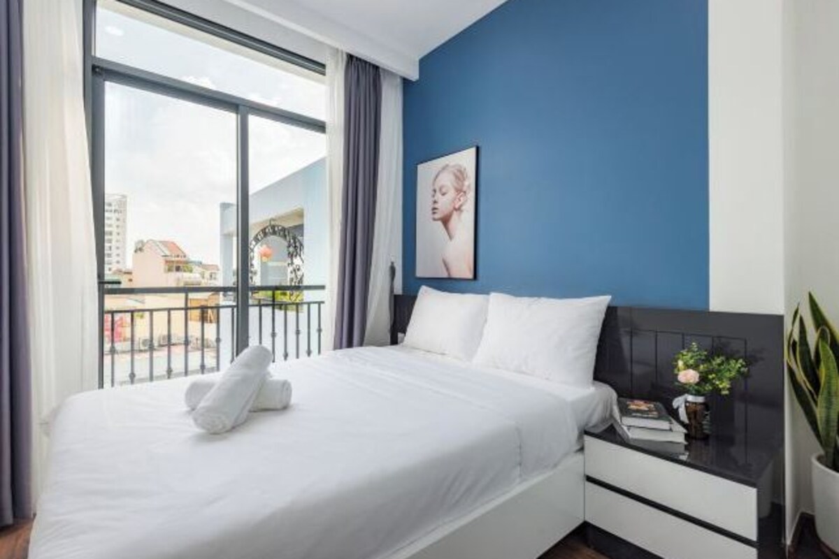 A modern bedroom with a large window, a bed with white linens, a nightstand with a book and a plant, and a blue accent wall.