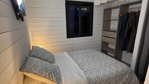 Un dormitorio pequeño con cama, mesita de noche, televisor montado en la pared, ventana y armario con ropa colgada.