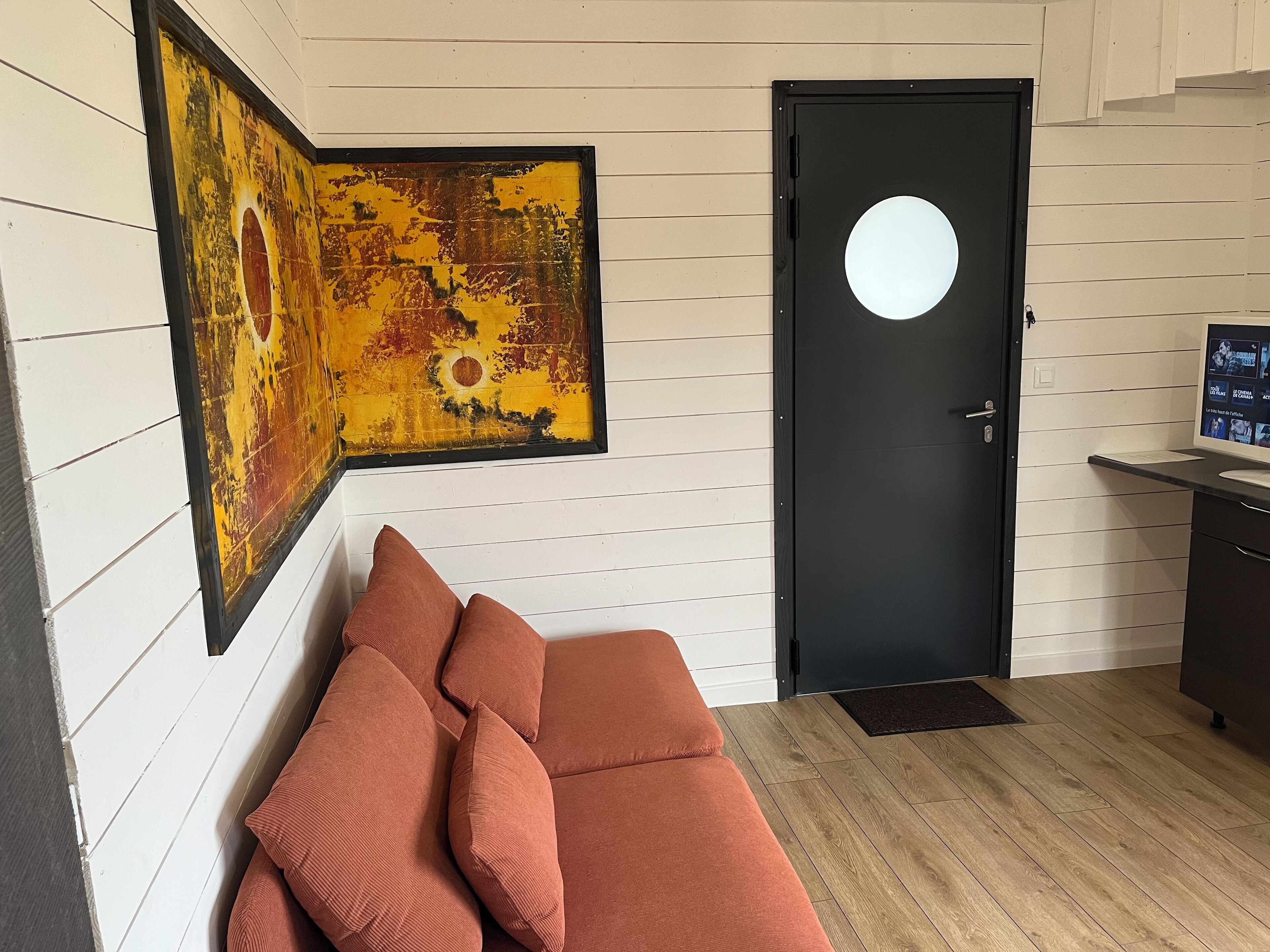 Una habitación con una puerta negra, un cuadro abstracto enmarcado, un sofá marrón con cojines y un escritorio pequeño con un monitor de computadora.
