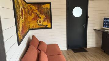 Una habitación con una puerta negra, un cuadro abstracto enmarcado, un sofá marrón con cojines y un escritorio pequeño con un monitor de computadora.