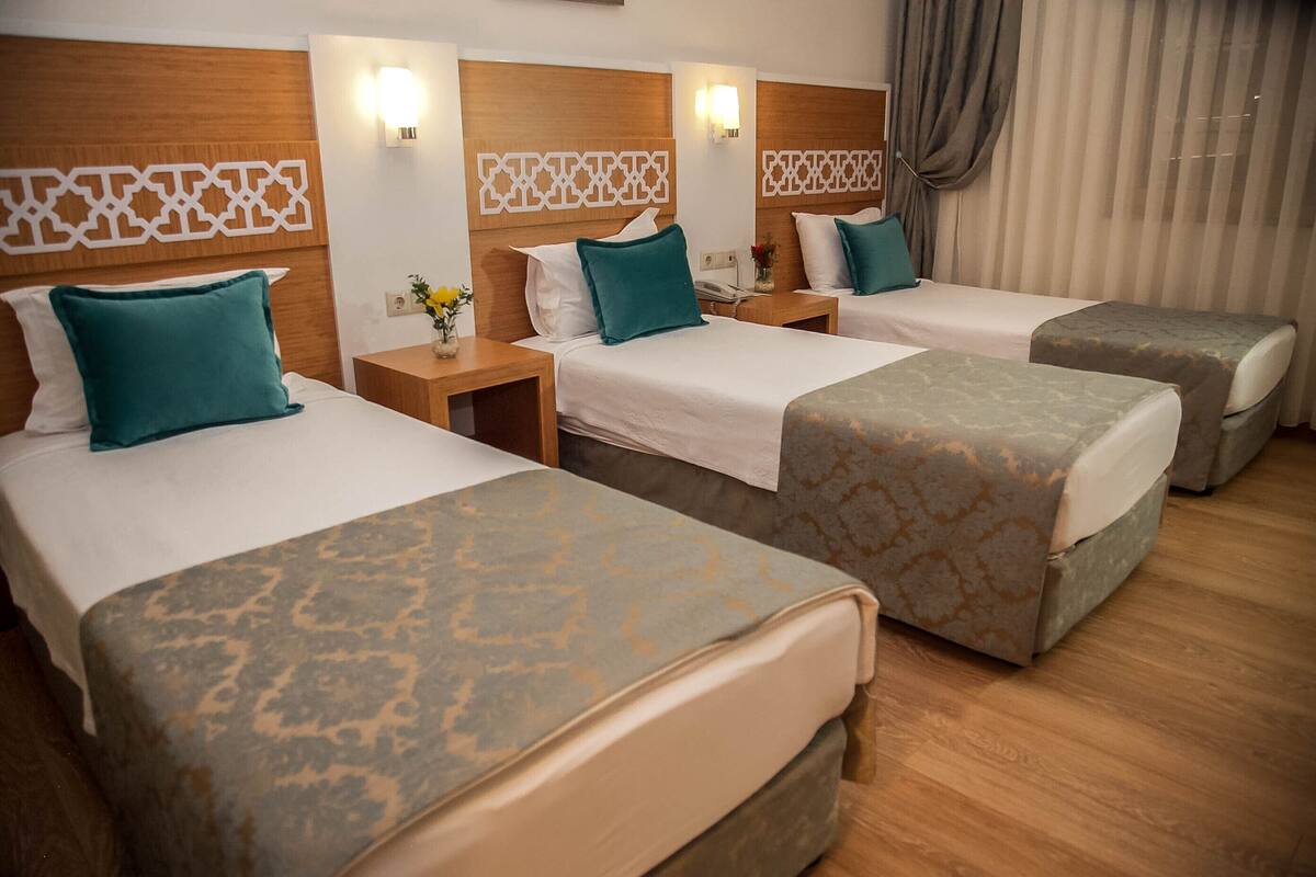 A hotel room with three single beds, each with a patterned bedspread and matching pillows. There are wooden headboards with decorative panels and wall-mounted lights.