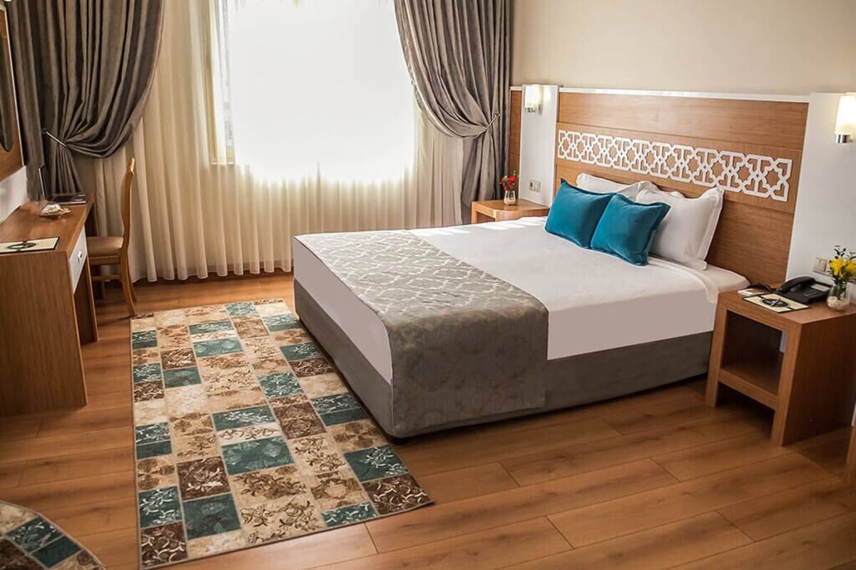 A hotel room with a large bed, a wooden headboard, a bedside table with a lamp, a nightstand, and a view of the outdoors through sheer curtains.