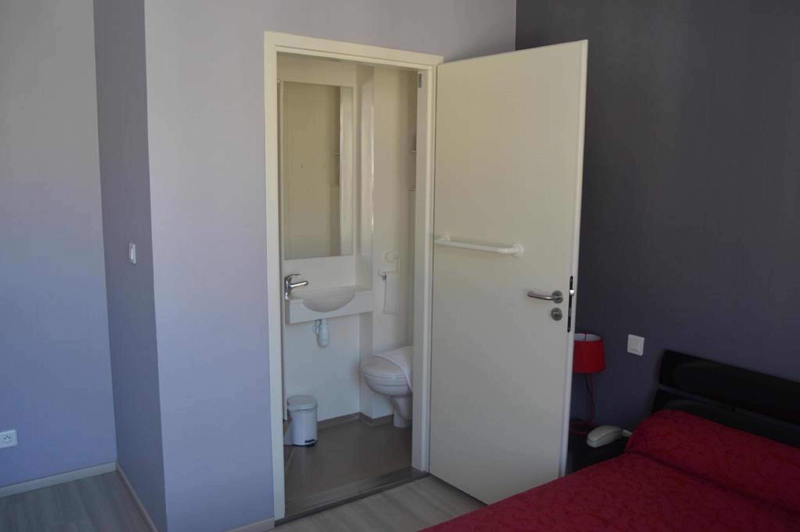 Une pièce avec un lit, une couverture rouge, une porte blanche donnant sur une salle de bain et une lampe rouge.