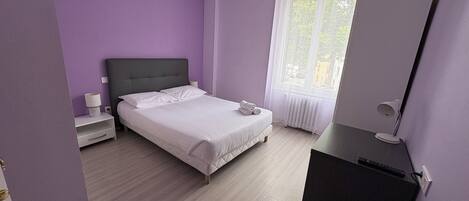 Een slaapkamer met een bed, nachtkastje, lamp en een bureau met stoel.