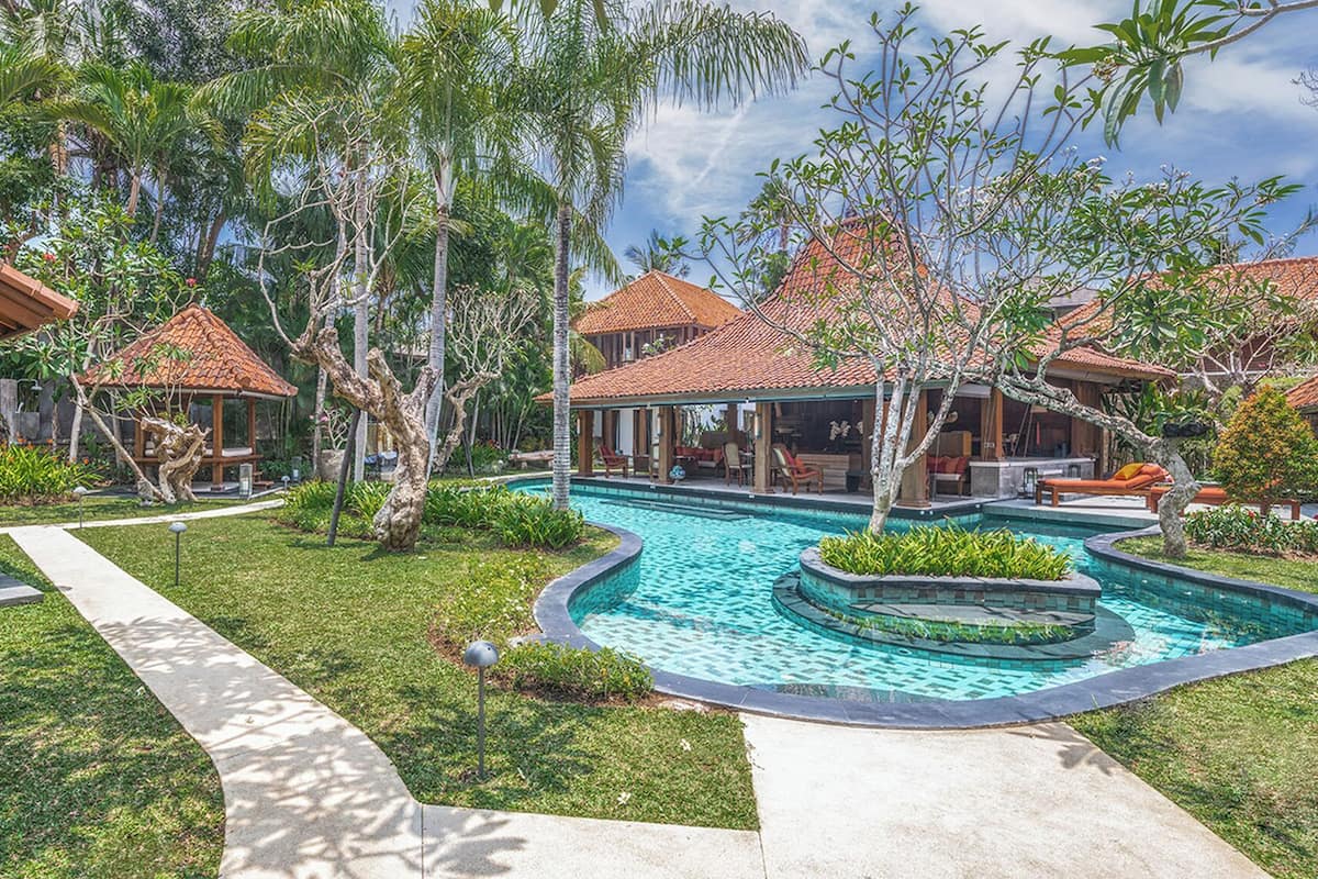 A resort with a central swimming pool, surrounded by palm trees and covered outdoor seating areas.