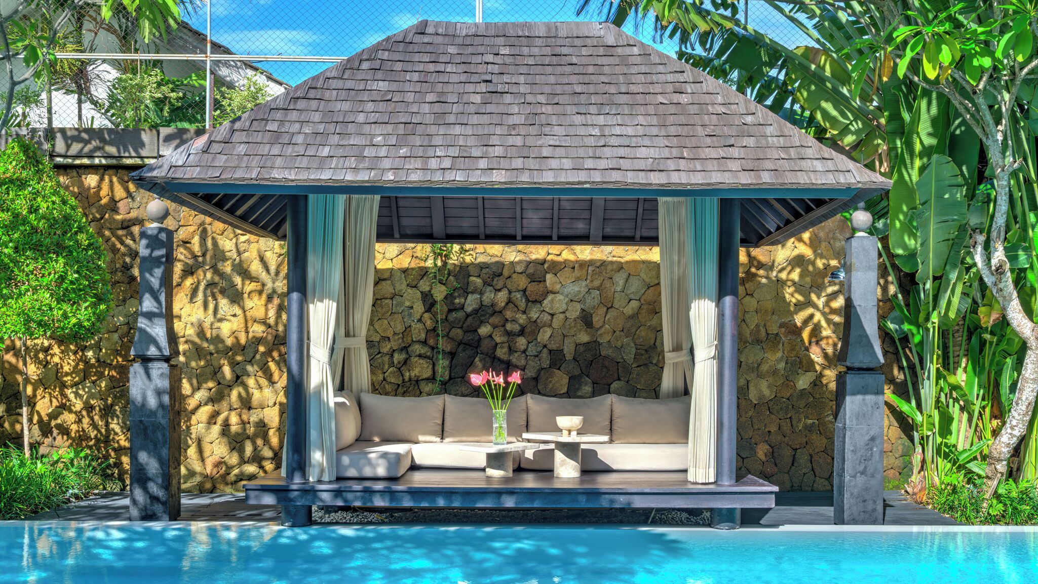 Un espace de détente abrité, avec un mur en pierre en arrière-plan, une piscine et une végétation tropicale.