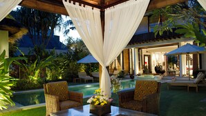 Zona piscina con mobili in vimini, un tavolo con composizione floreale e un’area relax coperta con tende bianche.