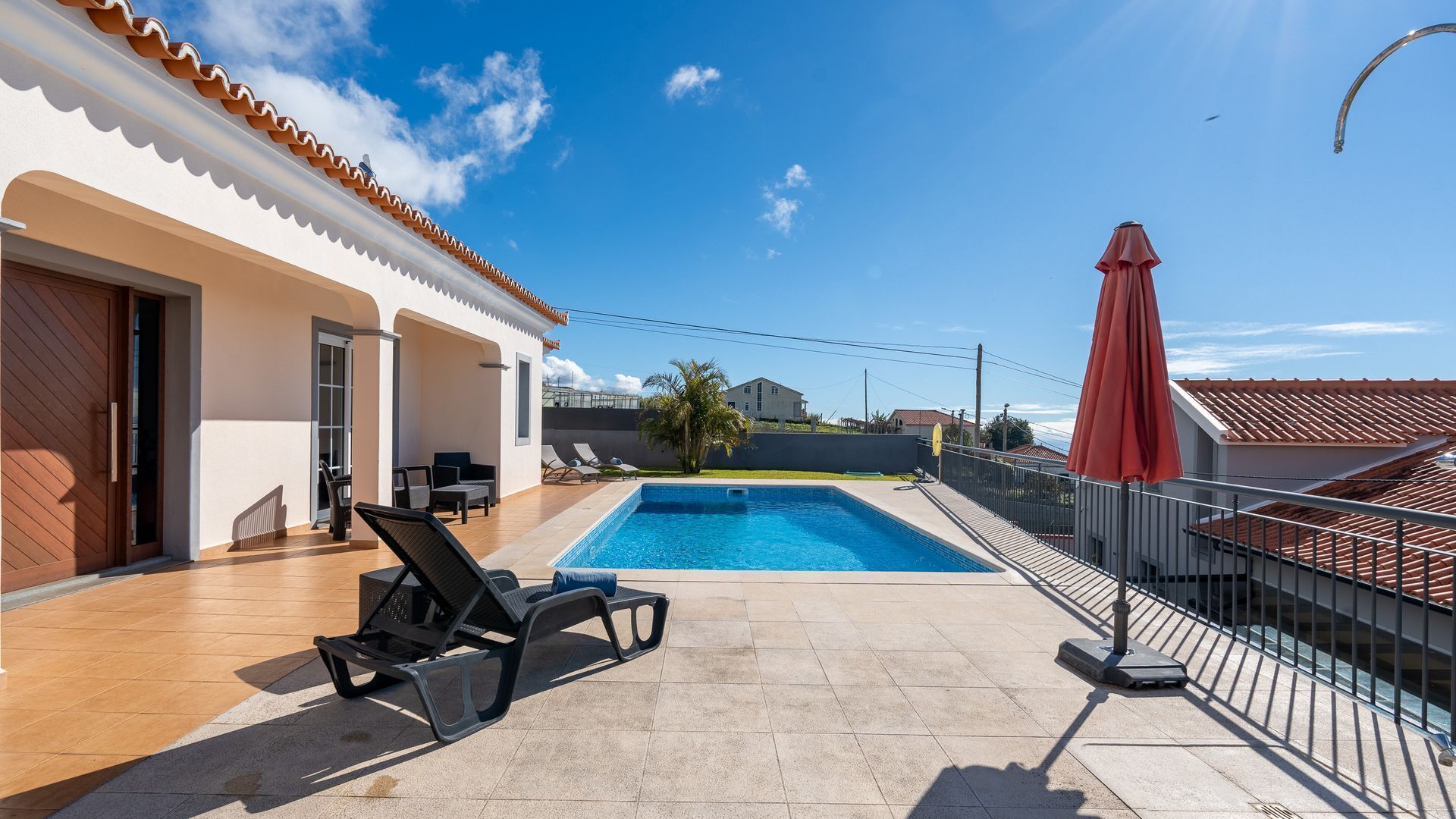 A swimming pool with lounge chairs, a red umbrella, and a view of a neighboring house.