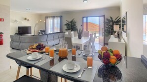 A modern living room with a grey sofa, a flat-screen TV, and a dining area with a table set for breakfast.