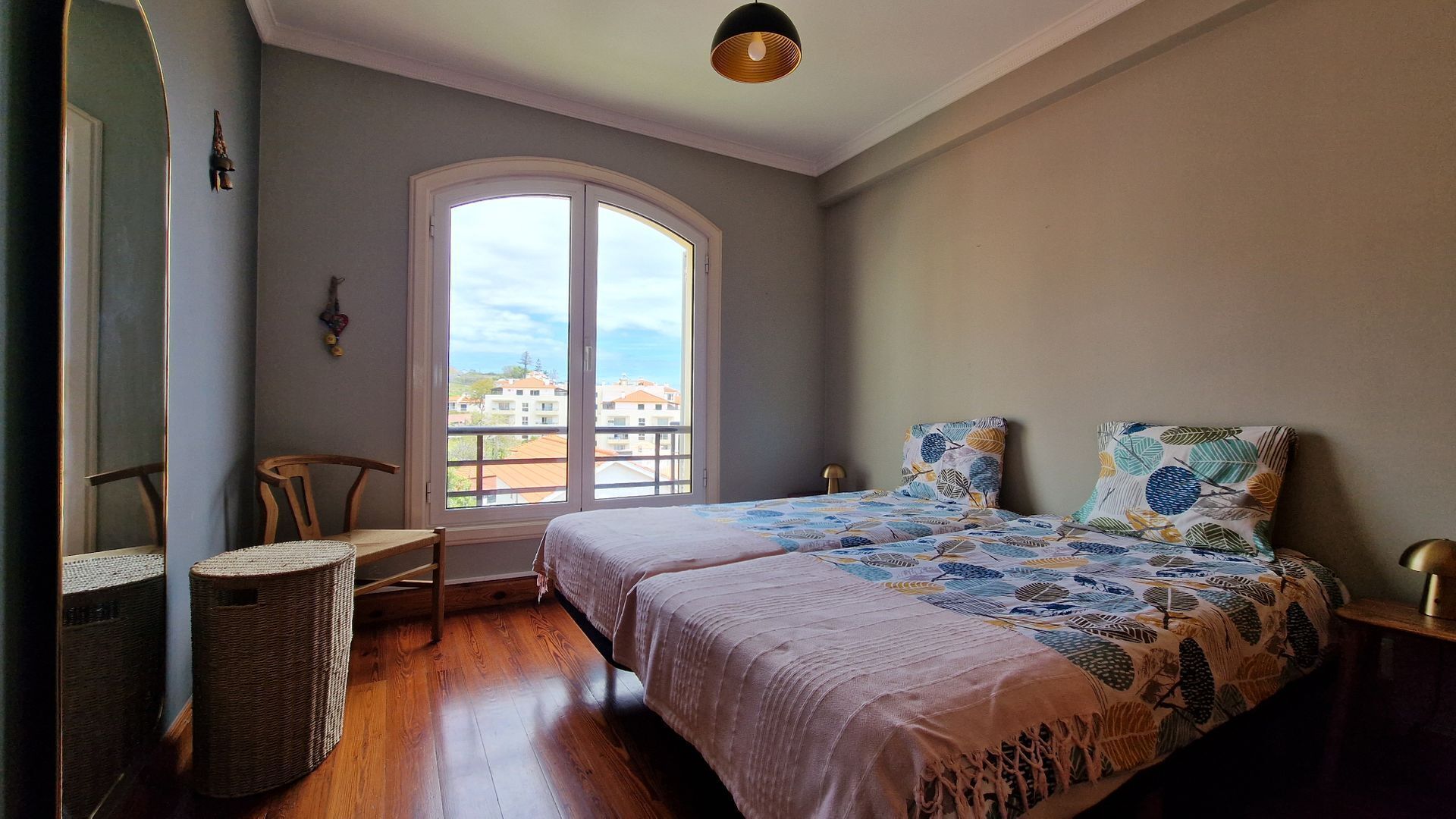 Ein Schlafzimmer mit großem Fenster, einem Bett mit gemusterten Bettbezügen, einem Sessel und einem kleinen Tisch.