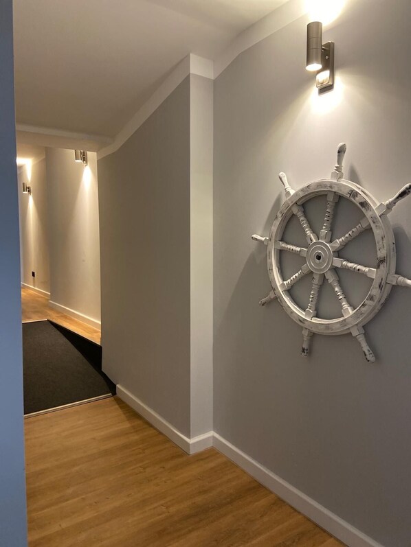 A hallway with a wooden floor, a grey wall, and a ship's wheel decoration.