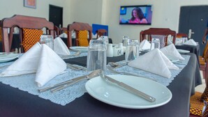A dining table set with white napkins, silverware, and plates, in a room with chairs and a television screen in the background.