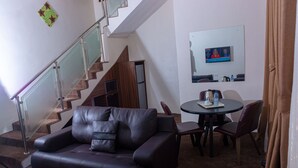 A living room with a leather sofa, a dining table with chairs, a television on the wall, and a staircase.