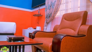 A room with a red-orange wall, a television mounted on the wall, a sofa, a small table, and a window with curtains. - Sweet Savor Hotels and Suites Annex (Lagos)