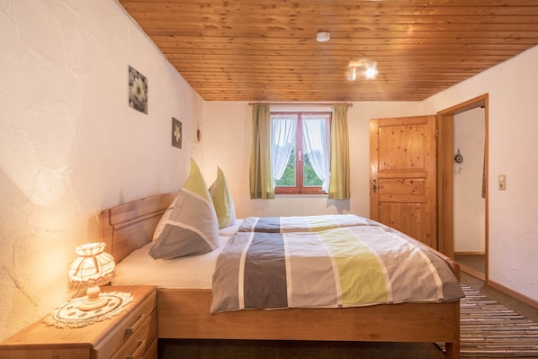 Une chambre avec un plafond en bois, une fenêtre avec des rideaux, un lit recouvert d’une couverture à rayures et une table de chevet avec une lampe.