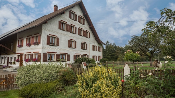 Ein mehrstöckiges Gebäude mit geneigtem Dach, zahlreichen Fenstern mit Fensterläden und einem gepflegten Garten mit verschiedenen Pflanzen und Blumen.