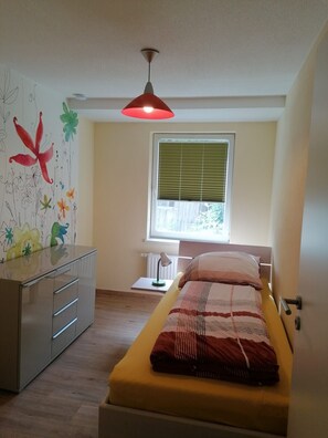 A bedroom with a bed, a desk, a radiator, a window with blinds, and a ceiling light.