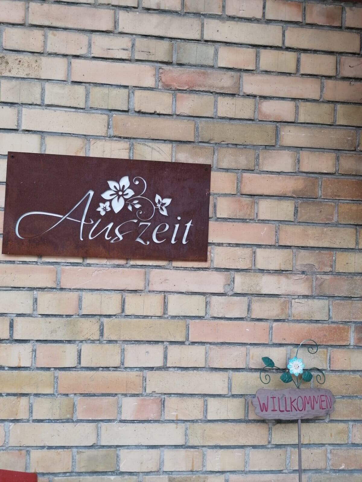 Eine Ziegelwand mit einem Schild, auf dem 'Auszeit' steht, und einem darunterliegenden Schild mit der Aufschrift 'WILLKOMMEN'.