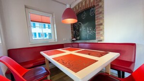 Ein Zimmer mit einem Tisch und vier roten Stühlen, einem Fenster mit Jalousien, einer Ziegelwand und einer Tafel mit Zeichnungen.