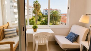A room with a view of buildings and palm trees, a white table with a chair, a sofa with a lamp, and a wooden bench.