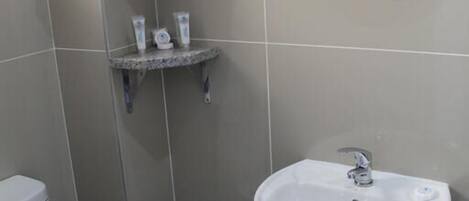 Un baño con inodoro, lavamanos, toallero con toallas dobladas, un estante pequeño con artículos de aseo personal y un espejo redondo.