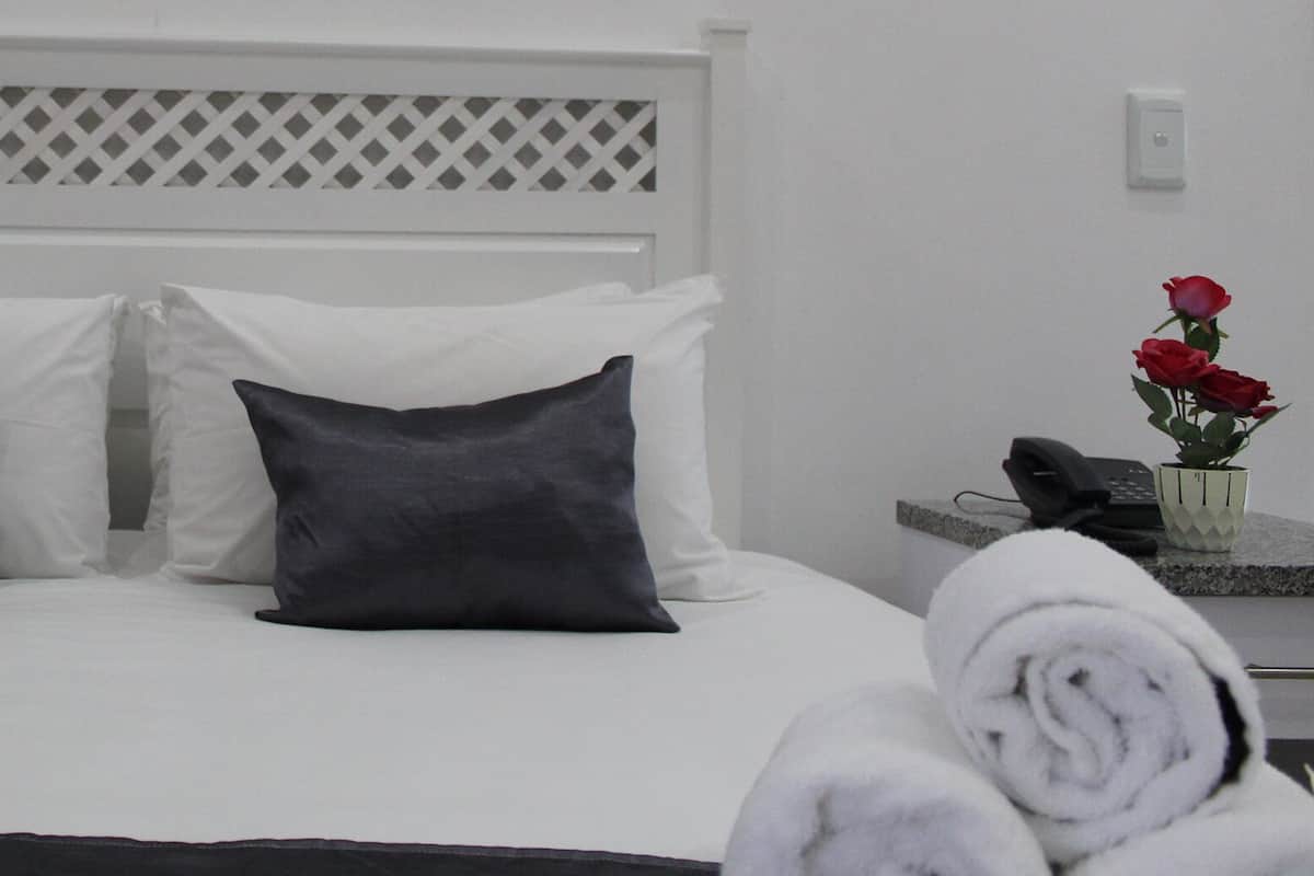 A hotel room with a bed, two pillows, a white headboard, a nightstand with a vase of red roses, and a rolled towel.