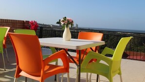Un balcon avec une table et des chaises blanches, un vase rempli de fleurs et une vue sur la mer.