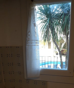 Une fenêtre ornée d’un rideau décoratif, donnant sur un palmier et une piscine.
