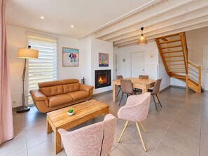 Ein Wohnzimmer mit Sofa, Couchtisch und Sesseln, einem Kamin, einem Essbereich mit Tisch und Stühlen sowie einer Treppe.