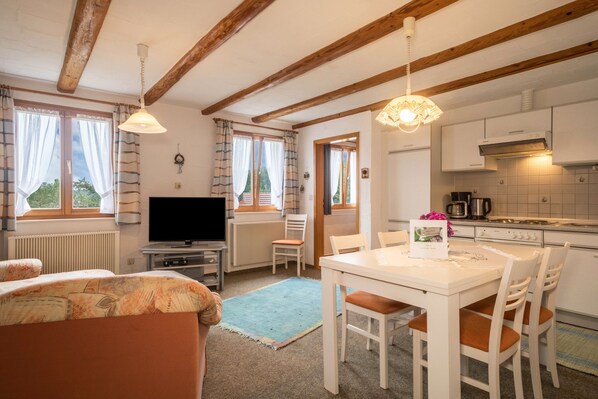 A room with a TV, a dining table with chairs, a kitchen area, and a window with curtains.