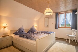A bedroom with a bed, bedside lamps, a chair, a window with curtains, and a wooden ceiling.