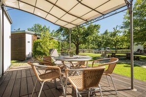 Eine überdachte Terrasse mit einem runden Tisch und Korbstühlen, eine kleine Gartenhütte und ein Rasen mit Bäumen.