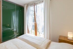 Un dormitorio con una cama grande, una mesita de noche con lámpara, un espejo y una ventana con cortinas traslúcidas.
