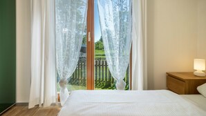 Ein Schlafzimmer mit Bett, Nachttisch mit Lampe und Blick auf einen Holzzaun und Grün durch transparente Vorhänge.