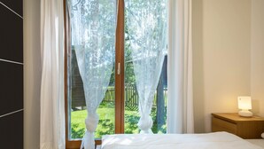 A bedroom with a bed, a nightstand with a lamp, and a view of a garden through a window with sheer curtains.