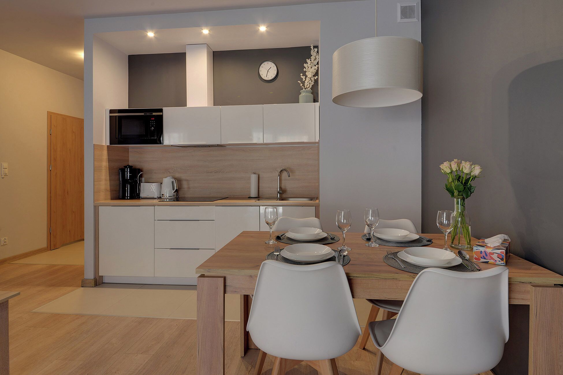 A modern kitchen with white cabinets, a microwave, and a sink. There is a dining table set with plates, glasses, and a vase of flowers.
