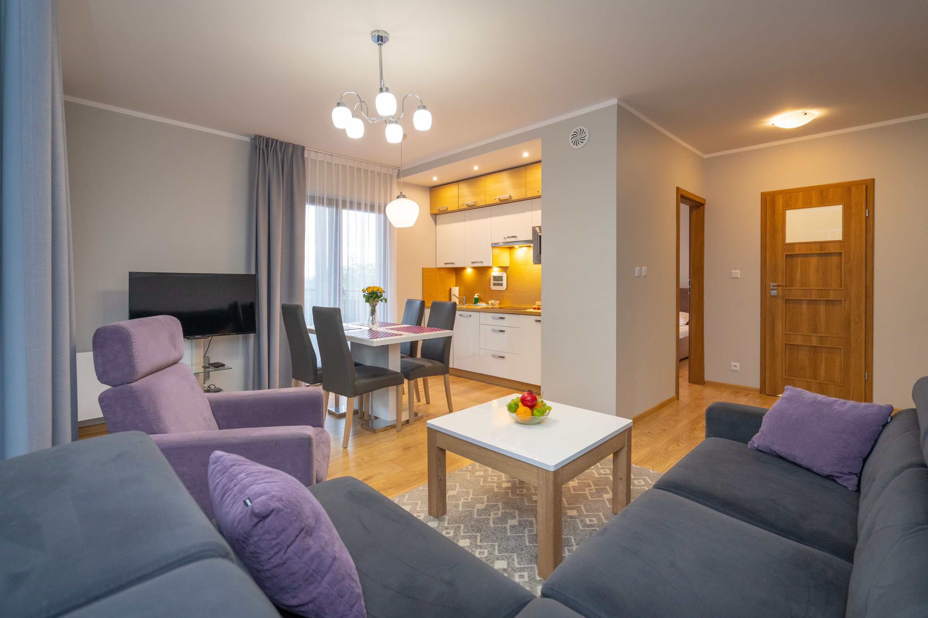 A modern living room with a grey sofa, a dining area with a table and chairs, a kitchen in the background, and a wooden door.