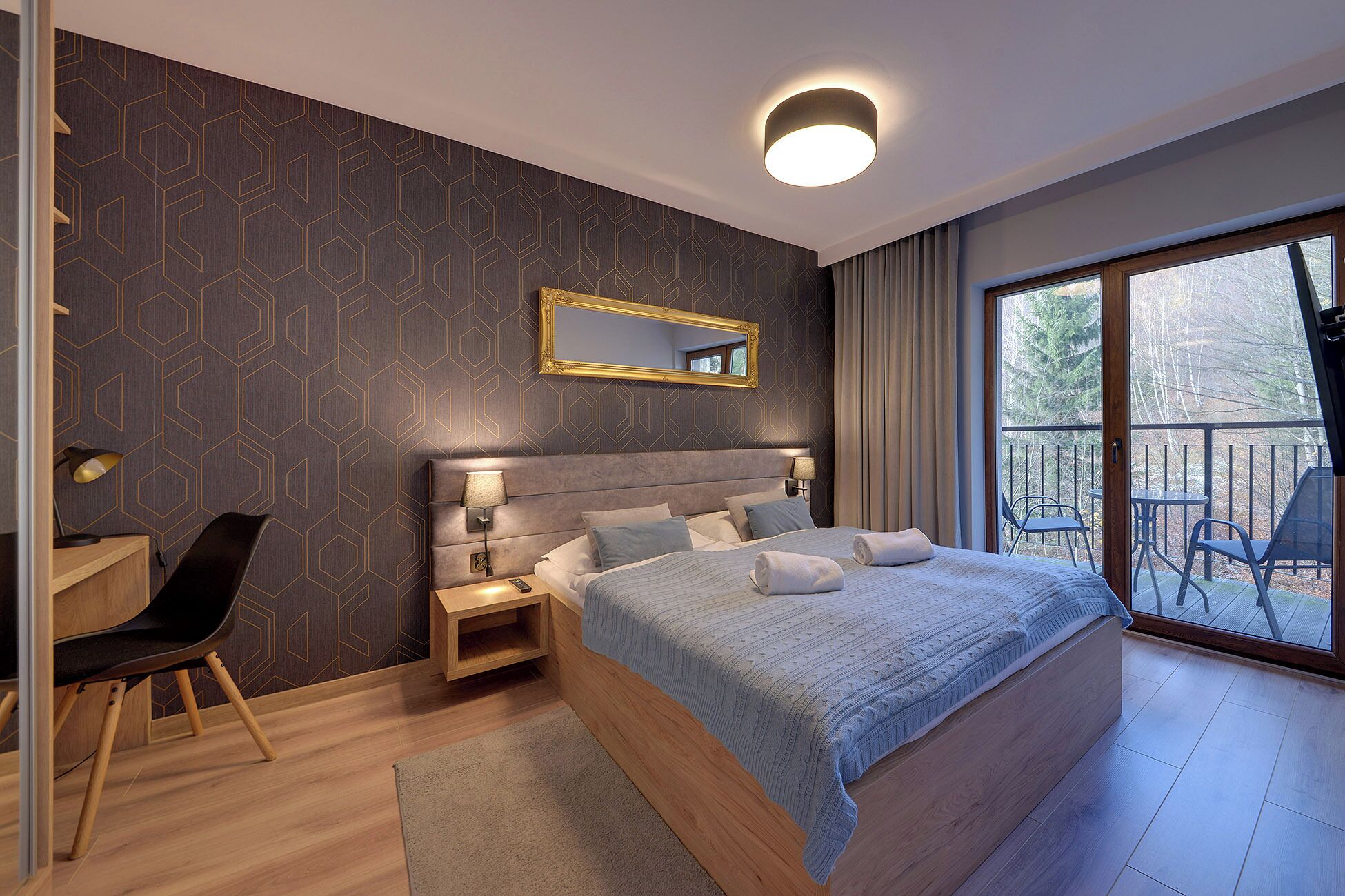 Een moderne slaapkamer met een groot bed, een nachtkastje, een bureau en een stoel. Er is een balkon met uitzicht op bomen.